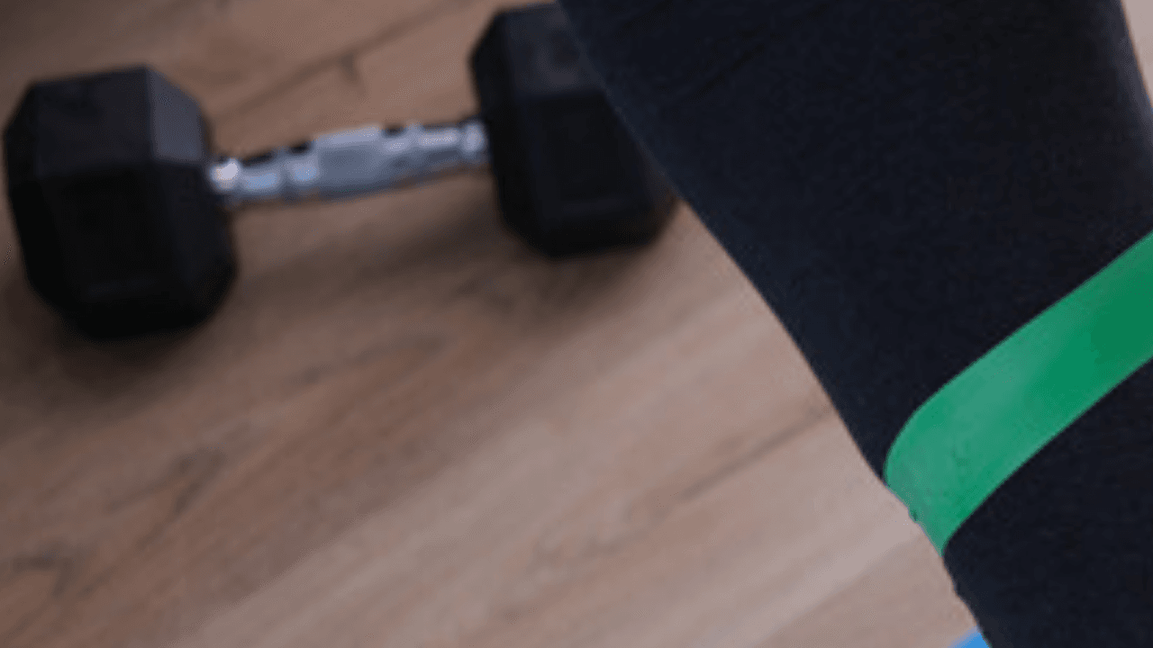 Träning med gummiband runt benen på matta hemma – hantel i bakgrunden